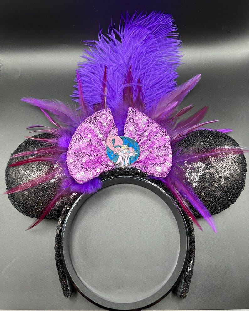 May include: A headband featuring two round ears covered in black sequins, a central light purple sequined bow, and a circular pin depicting a cartoon character. The design is accented with vibrant purple feathers extending upwards and outwards.