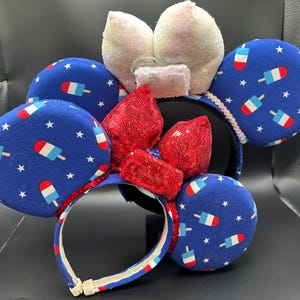 K&ouml;nnte beinhalten: Ein festliches Stirnband mit gro&szlig;en, runden Ohren, die mit blauem Stoff mit einem Eis am Stiel- und Sternmuster bedeckt sind. Das Stirnband hat ein rot-wei&szlig; gestreiftes Design mit einer roten Pailletten-Schleife und wei&szlig;en Pailletten-Ohren. Ein lustiges Accessoire.