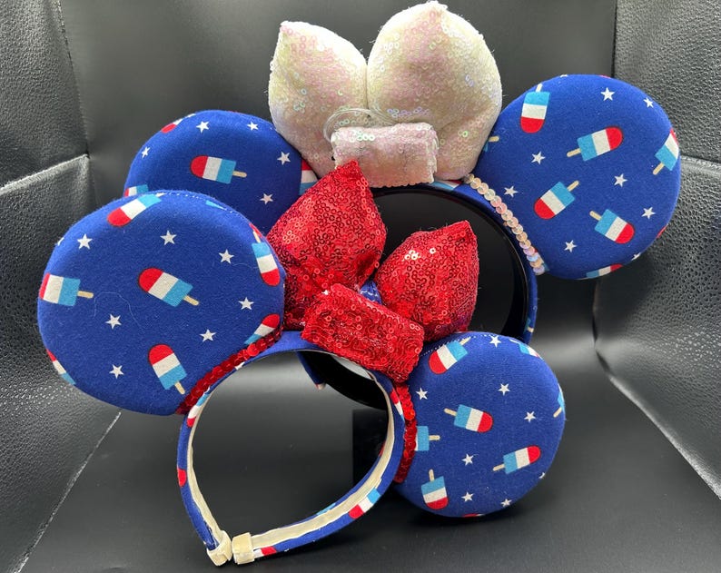 K&ouml;nnte beinhalten: Ein festliches Stirnband mit gro&szlig;en, runden Ohren, die mit blauem Stoff mit einem Eis am Stiel- und Sternmuster bedeckt sind. Das Stirnband hat ein rot-wei&szlig; gestreiftes Design mit einer roten Pailletten-Schleife und wei&szlig;en Pailletten-Ohren. Ein lustiges Accessoire.