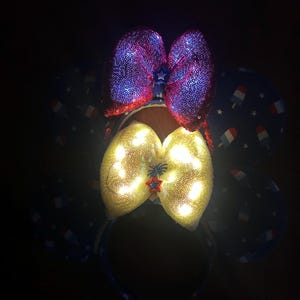 K&ouml;nnte beinhalten: Ein festliches Stirnband mit zwei Pailletten-Schleifen. Die obere Schleife ist rot und lila, w&auml;hrend die untere Schleife gelb mit eingebetteten Lichtern ist. Das Stirnband hat einen blauen Stoff mit Eis am Stiel- und Sternmustern.