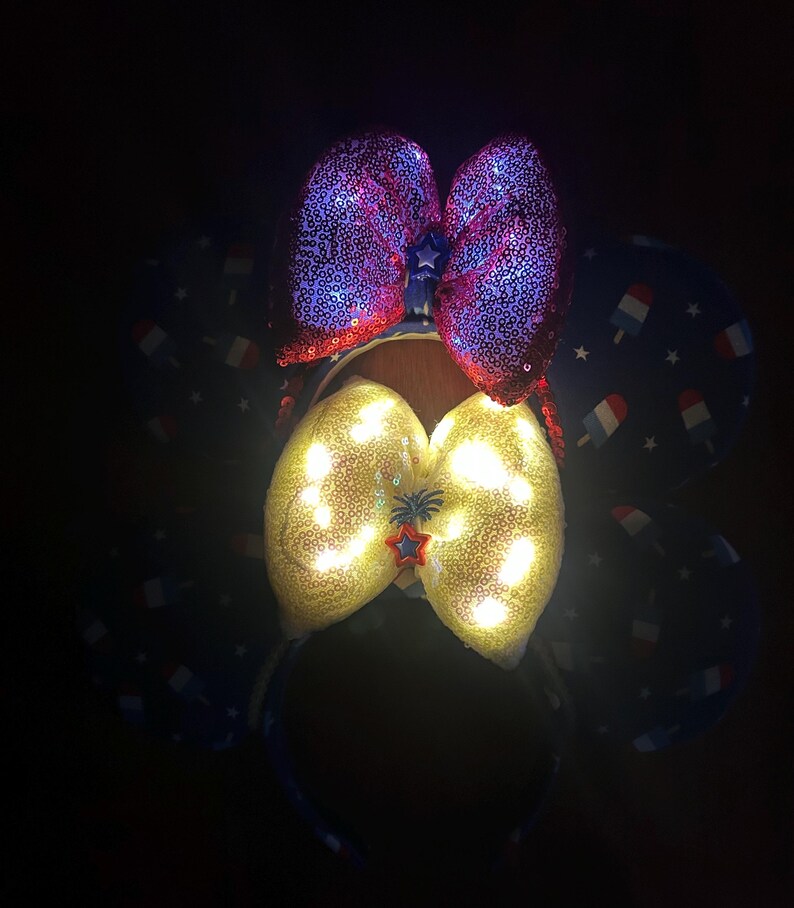 K&ouml;nnte beinhalten: Ein festliches Stirnband mit zwei Pailletten-Schleifen. Die obere Schleife ist rot und lila, w&auml;hrend die untere Schleife gelb mit eingebetteten Lichtern ist. Das Stirnband hat einen blauen Stoff mit Eis am Stiel- und Sternmustern.