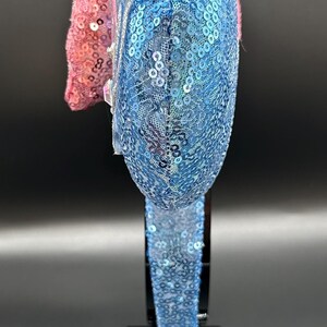 May include: A blue and pink sequined headband. The blue section is covered in small, iridescent sequins. The pink section is made of a textured, sequined fabric. The headband is displayed on a black stand.