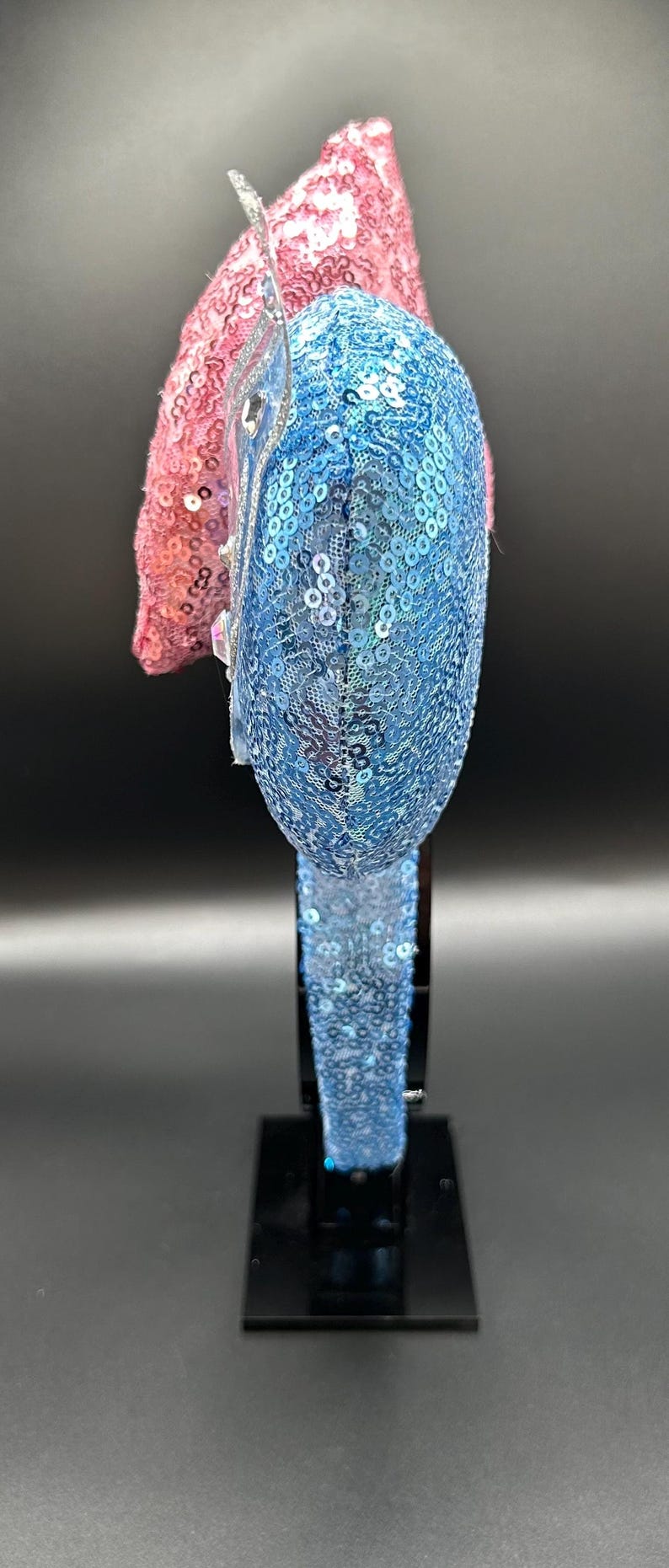 May include: A blue and pink sequined headband. The blue section is covered in small, iridescent sequins. The pink section is made of a textured, sequined fabric. The headband is displayed on a black stand.