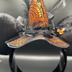 Puede incluir: Una diadema con dos grandes orejas de terciopelo negro y un sombrero de bruja central. El sombrero es naranja con encaje negro, plumas negras y una cinta negra con tachuelas plateadas. Una peque&ntilde;a gema naranja se encuentra en una de las orejas.