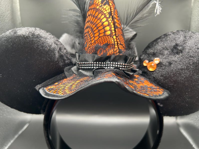 Puede incluir: Una diadema con dos grandes orejas de terciopelo negro y un sombrero de bruja central. El sombrero es naranja con encaje negro, plumas negras y una cinta negra con tachuelas plateadas. Una peque&ntilde;a gema naranja se encuentra en una de las orejas.