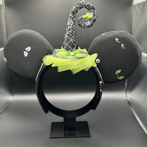 May include: Black Halloween-themed mouse ears with a witch hat design. The ears are black with white ghost-like shapes and green accents. The witch hat is black with a white spiderweb pattern and a green tulle brim. The headband is black.