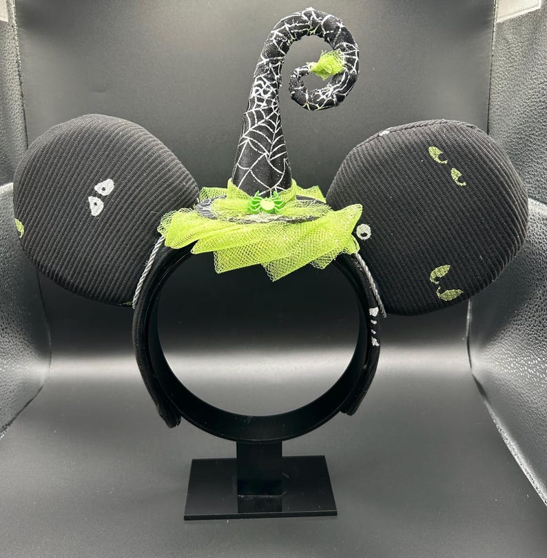 May include: Black Halloween-themed mouse ears with a witch hat design. The ears are black with white ghost-like shapes and green accents. The witch hat is black with a white spiderweb pattern and a green tulle brim. The headband is black.