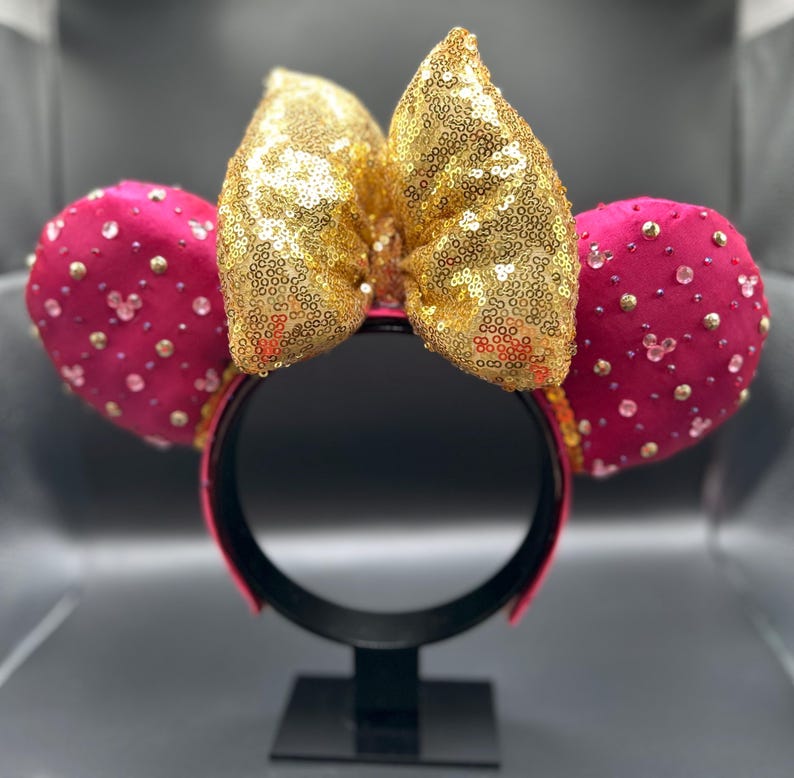 May include: A pair of mouse ears with a gold sequined bow and fuchsia ears. The ears are embellished with small, clear and gold-colored rhinestones. The headband is fuchsia and black.