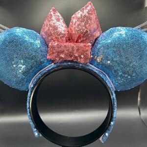 May include: A blue sequined headband with two round ears and a pink sequined bow. The headband has clear, wing-shaped accents on either side of the ears. The ears and bow are covered in sequins, creating a sparkling effect.