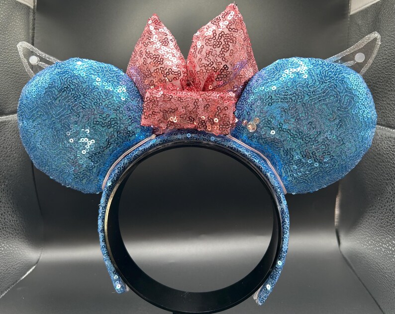 May include: A blue sequined headband with two round ears and a pink sequined bow. The headband has clear, wing-shaped accents on either side of the ears. The ears and bow are covered in sequins, creating a sparkling effect.
