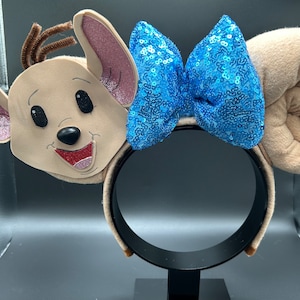 May include: A pair of beige mouse ears with a blue sequined bow. The ears are plush and have a 3D face on one ear. The headband is black.
