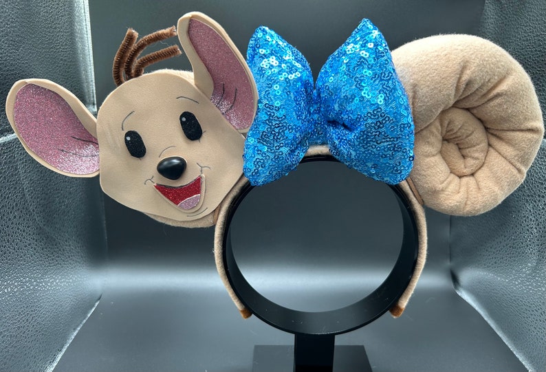 May include: A pair of beige mouse ears with a blue sequined bow. The ears are plush and have a 3D face on one ear. The headband is black.