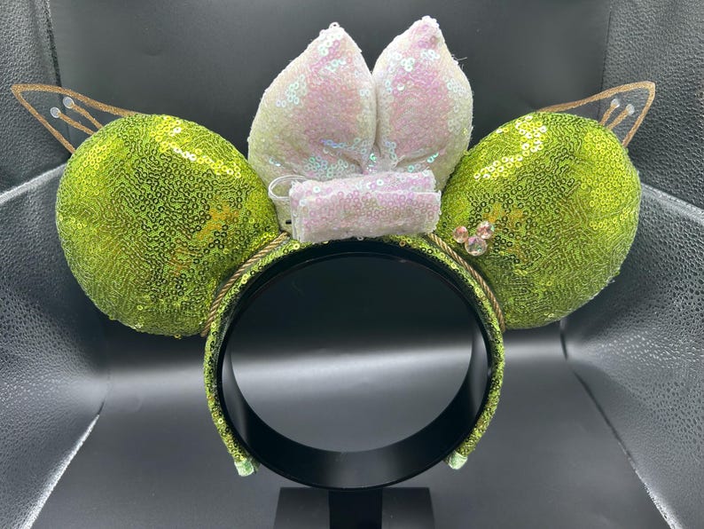 Op de afbeelding: Een haarband met twee grote, ronde, limoengroene oren bedekt met lovertjes. Tussen de oren zit een centrale strik van iriserende, lichtroze en witte lovertjes. Twee puntige, goudomrande, doorschijnende vleugels steken uit bovenop elk oor.