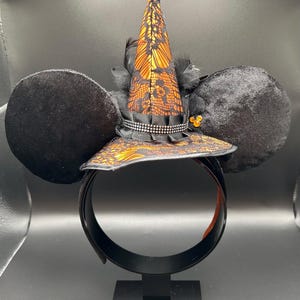 May include: Black mouse ear headband with a witch hat design. The hat is orange and black with lace detailing, a black ribbon, and a rhinestone band. Black feathers and a small gold embellishment add to the design.