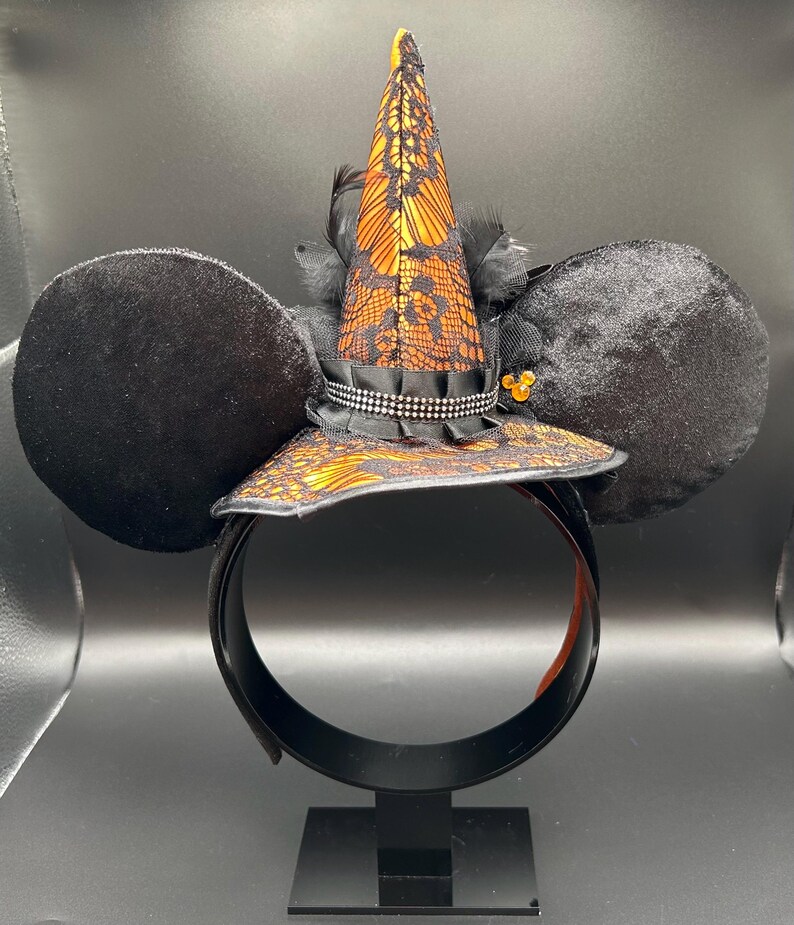 May include: Black mouse ear headband with a witch hat design. The hat is orange and black with lace detailing, a black ribbon, and a rhinestone band. Black feathers and a small gold embellishment add to the design.
