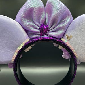 Puede incluir: Una diadema con dos orejas grandes, suaves y de color morado claro, con un lazo central iridiscente del mismo tono. La base de la diadema est&aacute; cubierta de lentejuelas moradas oscuras, con un peque&ntilde;o detalle de lentejuelas en el centro.