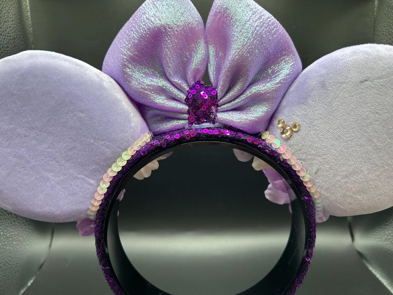 Puede incluir: Una diadema con dos orejas grandes, suaves y de color morado claro, con un lazo central iridiscente del mismo tono. La base de la diadema est&aacute; cubierta de lentejuelas moradas oscuras, con un peque&ntilde;o detalle de lentejuelas en el centro.