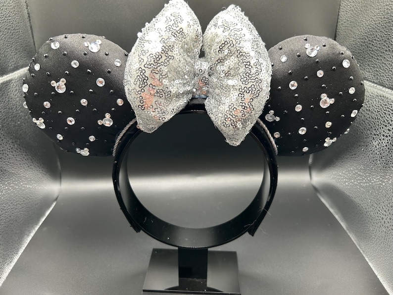 May include: Black headband with two round black ears adorned with clear rhinestones and small white Mickey Mouse silhouettes. A silver sequined bow sits between the ears. The headband is on a black stand.