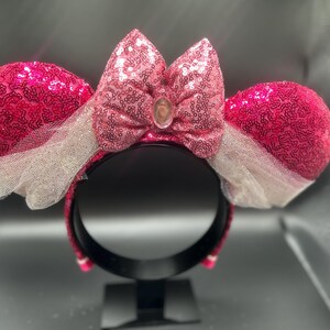 Op de afbeelding: Roze en witte Minnie Mouse oren met pailletten, met een roze strik en een heldere strasssteen in het midden.
