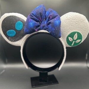 May include: A pair of white sequined mouse ears with a black and teal design on one ear and a green leaf emblem on the other. A galaxy-print bow sits atop the headband.