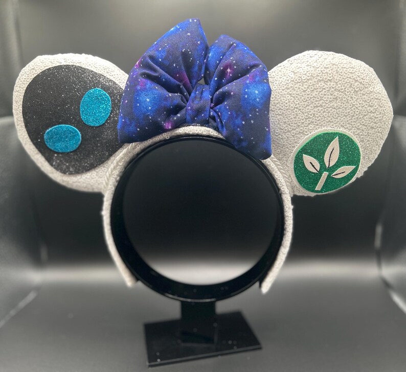 May include: A pair of white sequined mouse ears with a black and teal design on one ear and a green leaf emblem on the other. A galaxy-print bow sits atop the headband.