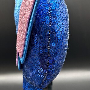 May include: A blue sequined headband with a pink glitter accent. The headband has a rounded shape and is decorated with small, shiny blue sequins. The pink accent is a rectangular shape. The headband is on a black stand.