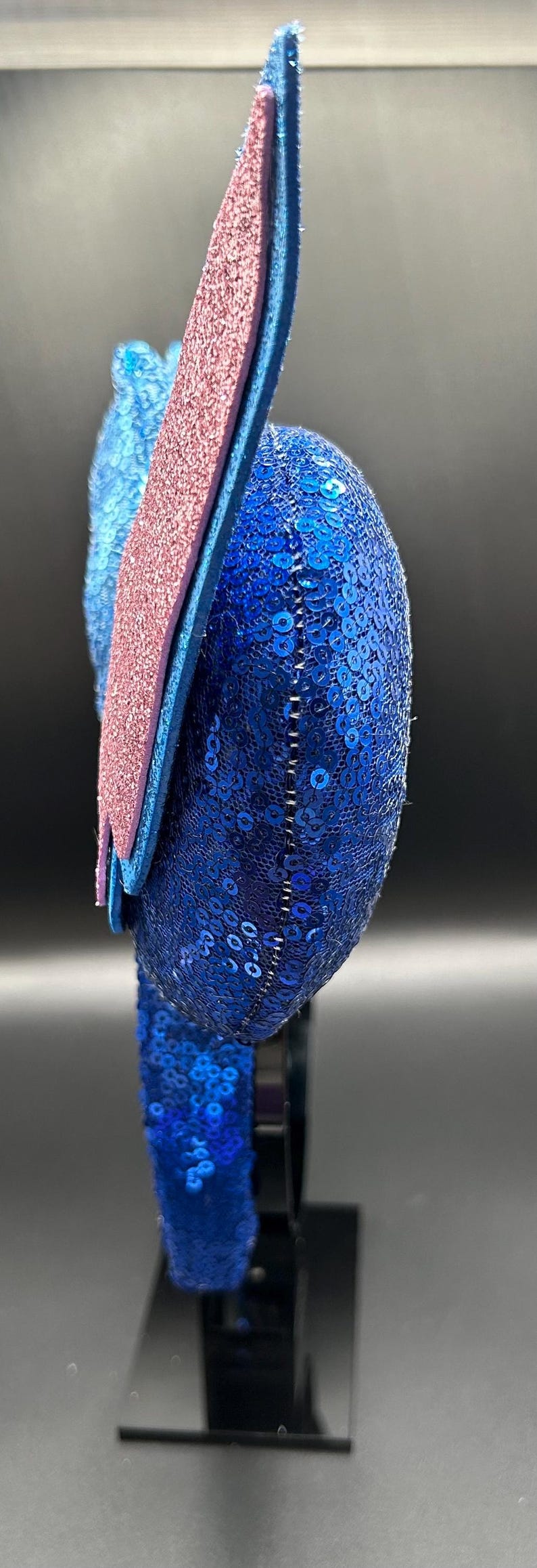 May include: A blue sequined headband with a pink glitter accent. The headband has a rounded shape and is decorated with small, shiny blue sequins. The pink accent is a rectangular shape. The headband is on a black stand.