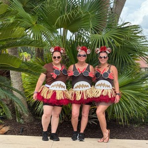 May include: Three individuals wearing brown tank tops and t-shirts decorated with black circular accents, red trim, and white jagged-edge details. Each outfit includes a layered skirt featuring a tan raffia fringe over a dark red tulle base.