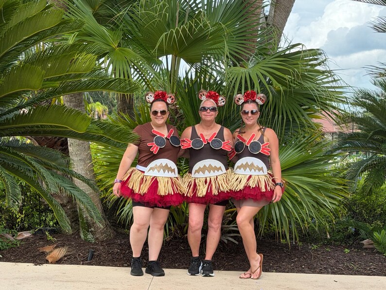 May include: Three individuals wearing brown tank tops and t-shirts decorated with black circular accents, red trim, and white jagged-edge details. Each outfit includes a layered skirt featuring a tan raffia fringe over a dark red tulle base.