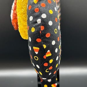 May include: A Halloween-themed headband with a black fabric band featuring white, orange, and yellow polka dots. The headband has a yellow sequined decoration and a red and white striped accent.