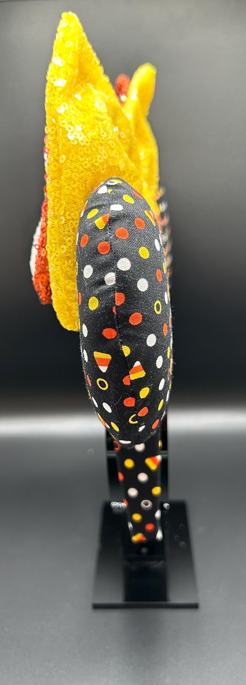 May include: A Halloween-themed headband with a black fabric band featuring white, orange, and yellow polka dots. The headband has a yellow sequined decoration and a red and white striped accent.
