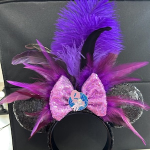 May include: A headband featuring two black sequined ears, a central pink sequined bow, and a circular enamel pin depicting a cartoon character. The accessory is adorned with an arrangement of vibrant purple and black feathers extending outward.
