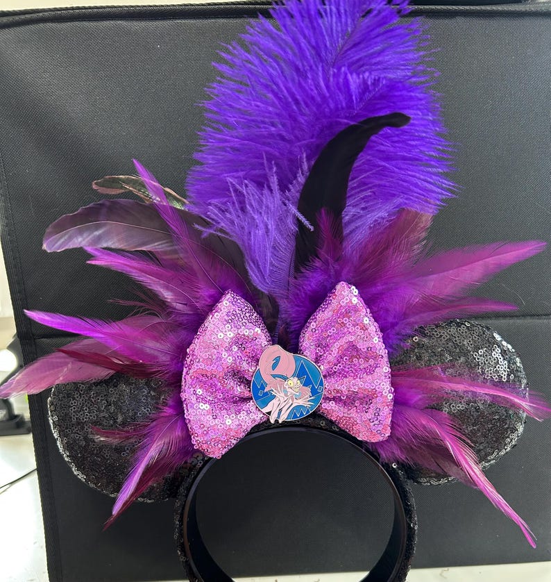 May include: A headband featuring two black sequined ears, a central pink sequined bow, and a circular enamel pin depicting a cartoon character. The accessory is adorned with an arrangement of vibrant purple and black feathers extending outward.