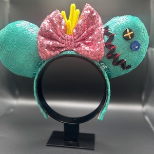 May include: Teal sequined mouse ears with a pink sequined bow and yellow accents. One ear has black stitching, a blue button, and a yellow cross. The black headband has teal trim. A fun accessory for themed events.