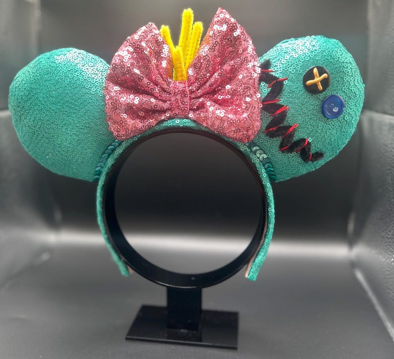 May include: Teal sequined mouse ears with a pink sequined bow and yellow accents. One ear has black stitching, a blue button, and a yellow cross. The black headband has teal trim. A fun accessory for themed events.