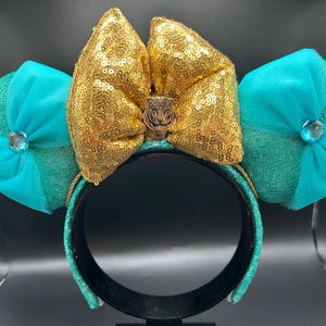 May include: A decorative headband with a gold sequined bow and tiger emblem, with turquoise sequined ears and turquoise bows, and a central jewel. The headband is black with turquoise accents.