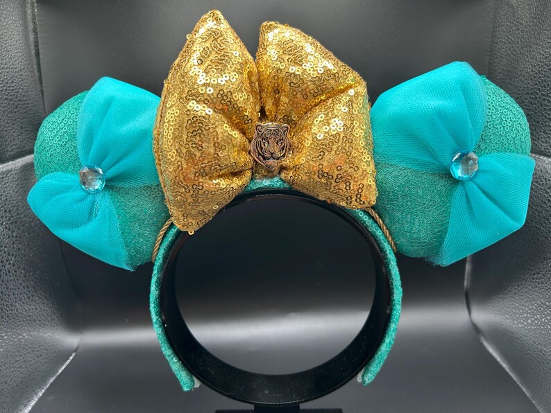 May include: A decorative headband with a gold sequined bow and tiger emblem, with turquoise sequined ears and turquoise bows, and a central jewel. The headband is black with turquoise accents.