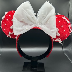 May include: Red and white mouse ear headband with a large white bow adorned with small pearls. The red ears are quilted and decorated with white bows and pearls. The headband is red and has a black stand.
