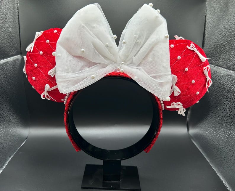May include: Red and white mouse ear headband with a large white bow adorned with small pearls. The red ears are quilted and decorated with white bows and pearls. The headband is red and has a black stand.