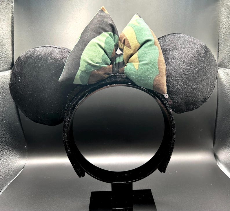 May include: Black sequined headband with two black plush ears and a camouflage bow. The bow features green, brown, and black patterns. The headband is supported by a black stand.