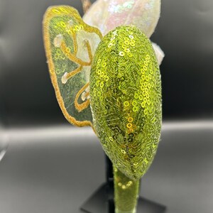 Op de afbeelding: Een haarband met een limoengroen oor vol lovertjes en een doorschijnende vleugel met goud geborduurde randen. Achter de vleugel is een wit iriserend element met lovertjes zichtbaar. De haarband is volledig bedekt met bijpassende limoengroene lovertjes.