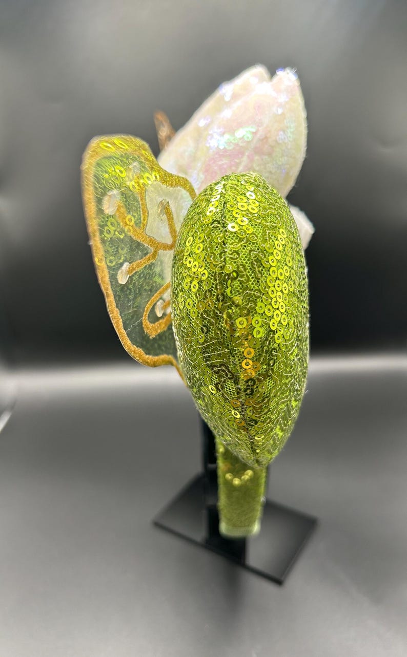 Op de afbeelding: Een haarband met een limoengroen oor vol lovertjes en een doorschijnende vleugel met goud geborduurde randen. Achter de vleugel is een wit iriserend element met lovertjes zichtbaar. De haarband is volledig bedekt met bijpassende limoengroene lovertjes.