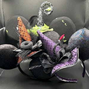 May include: A collection of Halloween-themed headbands featuring black, purple, and orange witch hats, bows, and decorative elements. The headbands have black ears with various textures, including velvet and sequin. Some headbands have a witch hat design.