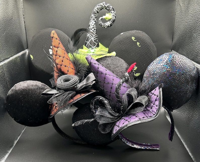 May include: A collection of Halloween-themed headbands featuring black, purple, and orange witch hats, bows, and decorative elements. The headbands have black ears with various textures, including velvet and sequin. Some headbands have a witch hat design.