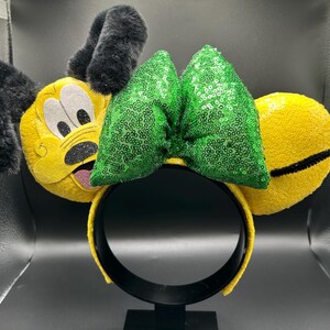 May include: A yellow headband with plush Pluto ears, a green sequined bow, and a black headband. The Pluto ears have black fur and a cartoon face. The bow is made of green sequins.