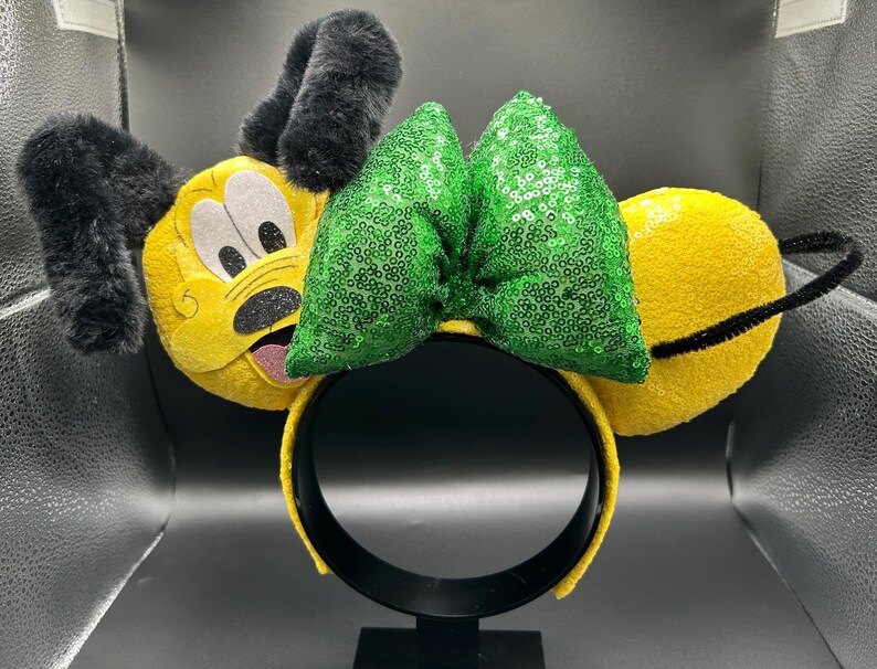 May include: A yellow headband with plush Pluto ears, a green sequined bow, and a black headband. The Pluto ears have black fur and a cartoon face. The bow is made of green sequins.