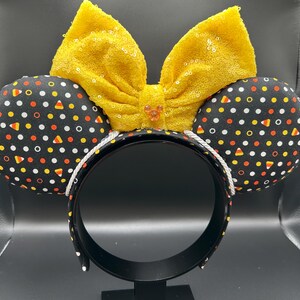 May include: Halloween-themed headband with black ears patterned with white dots, orange, yellow, and candy corn shapes. A large, yellow sequined bow sits atop the headband. The headband is black with a white trim.