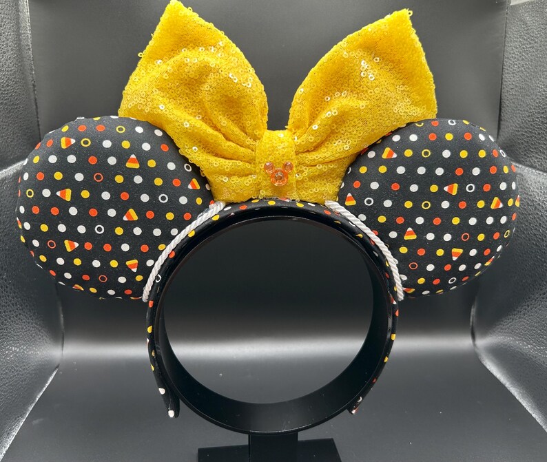 May include: Halloween-themed headband with black ears patterned with white dots, orange, yellow, and candy corn shapes. A large, yellow sequined bow sits atop the headband. The headband is black with a white trim.