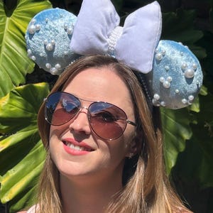 May include: A light blue headband with two round ears covered in faux pearls. A large white bow with pearl accents sits between the ears. The headband is worn by a person wearing sunglasses.
