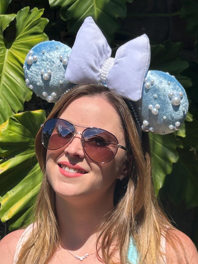 May include: A light blue headband with two round ears covered in faux pearls. A large white bow with pearl accents sits between the ears. The headband is worn by a person wearing sunglasses.